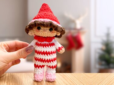 Gehäkelter Amigurumi-Wichtel mit roter Mütze und rot-weiß gestreiftem Anzug, von einer Hand gehalten, unscharfer Weihnachts­hintergrund.