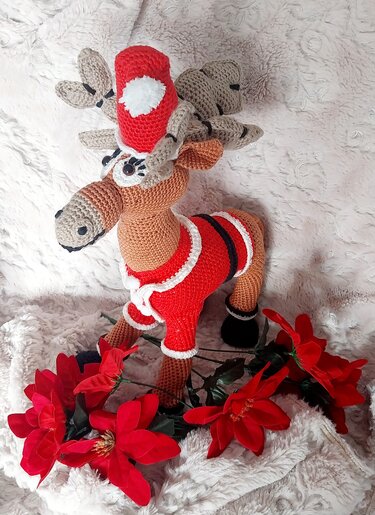 Gehäkeltes Amigurumi-Rentier im Weihnachtsmann-Outfit mit roter Mütze und großem Geweih, vor roten Weihnachtssternen
