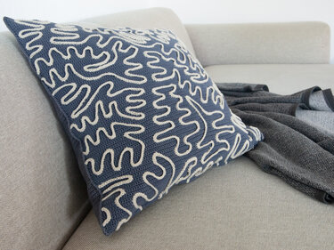 Square blue crocheted pillow with off-white abstract squiggle pattern on a light beige sofa.