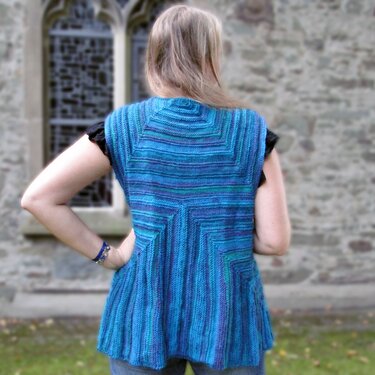 Back view of a woman wearing a blue, purple, and green variegated knitted sleeveless vest with a geometric back pattern.