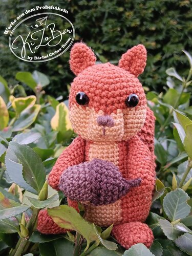 Gehäkeltes Amigurumi-Eichhörnchen (rostrot) sitzt in Blättern und hält eine gehäkelte Nuss