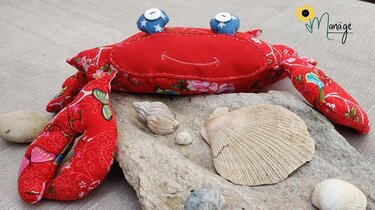 Genähte Stoffkrabbe aus rotem Musterstoff mit Knopfaugen, auf einem Stein neben Muscheln.