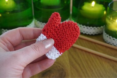Hand hält kleines gestricktes rotes Herz mit weißem Rand vor grünen Kerzen und Stricknadeln