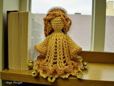 Gehäkeltes Engel-Amigurumi in senfgelbem Garn mit Rüschenrock und goldenen Glöckchen auf Fensterbank neben Buch