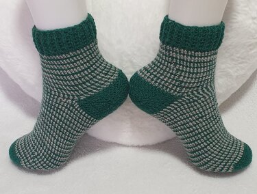Paar gehäkelter Knöchelsocken in Grün und Hellgrau mit gemustertem Schaft und einfarbigen Bündchen, Fersen und Spitzen auf weißem Untergrund.