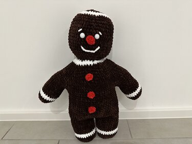 Gehäkelter dunkelbrauner Lebkuchenmann Amigurumi mit weißen Zierkanten und roten Knöpfen, frontal vor weißer Wand