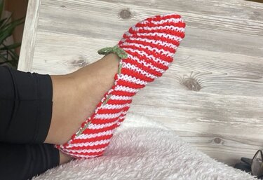 Fuß mit gestricktem rot-weiß gestreiftem Slipper und grüner Schleife auf weißem, flauschigem Untergrund.