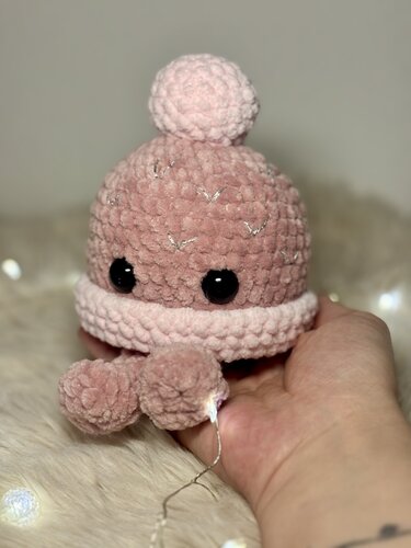 Rosa gehäkelter Amigurumi-Oktopus mit schwarzen Sicherheitsaugen und Pompons, in einer Hand auf flauschigem Untergrund vor Lichterkette gehalten.