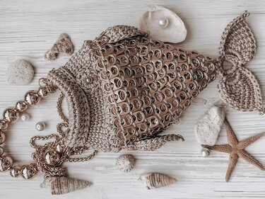 Beige hand-crocheted fish bag with ring-pull tab scales and beaded-chain strap on a light wooden surface with shells