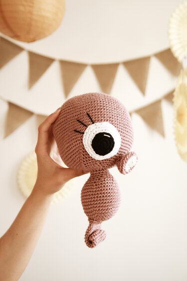 Hand hält rosabraune Amigurumi-Figur mit großem Auge und spiraligem unteren Ende, im Hintergrund beige Wimpelketten.