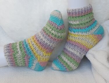 Handgehäkelte Socken mit pastellfarbenen Streifen auf weißem, flauschigem Hintergrund.