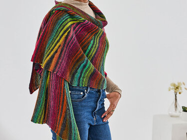 Gestricktes Tuch in bunten Regenbogenfarben, locker um Schultern drapiert über beigem Oberteil und Jeans.