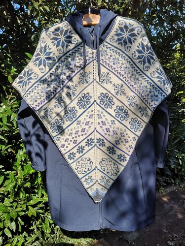 Gestrickter, spitz zulaufender Damenponcho mit blau-beigefarbenem Ornament- und Sternmuster, auf einem Kleiderbügel über einer dunklen Jacke aufgehängt.