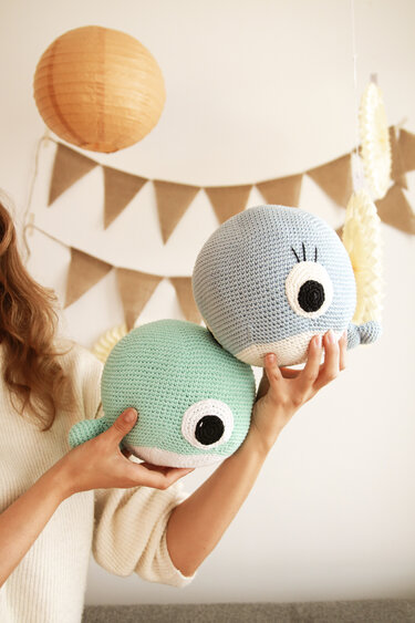 Zwei handgehäkelte Wal-Amigurumi in Hellblau und Mintgrün, in Händen gehalten; im Hintergrund Wimpelkette und Papierlaterne.