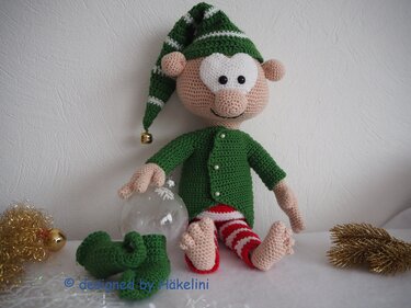 Gehäkelter Weihnachtself in Grün, Rot und Weiß mit Zipfelmütze, Jacke und gestreiften Hosen, umgeben von Weihnachtsdekoration.