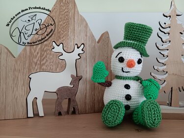 Gehäkelter Amigurumi-Schneemann mit grünem Zylinder, grünen Handschuhen und grünen Schuhen vor Holzfiguren von Rentier und Tannenbaum