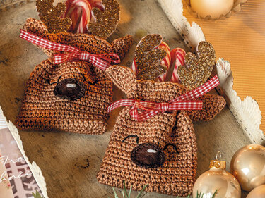 Zwei braune gehäkelte Rentier-Säckchen mit karierten Schleifen und glitzernden Geweihen, gefüllt mit Zuckerstangen auf einem Holztablett.