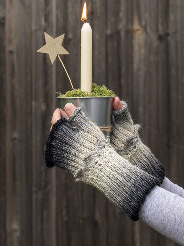 Hände mit gestrickten fingerlosen Armstulpen im Ombre‑Grau halten einen kleinen Topf mit brennender Kerze