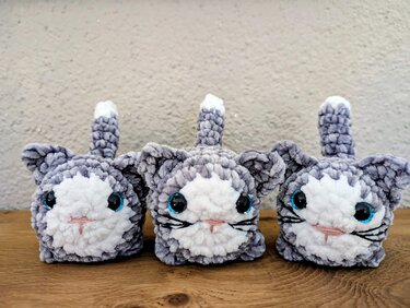 Drei gehäkelte Amigurumi-Katzen in Grau und Weiß auf Holzuntergrund vor strukturierter Wand.