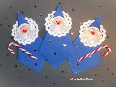 Drei blaue Wichtelfiguren mit gehäkelten Spitzenkragen, roten Nasen und Zuckerstangen auf grauem Hintergrund