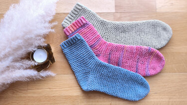 Drei gehäkelte Socken in Beige, Rosa und Blau auf Holzbrett neben Kerze und Pampasgras