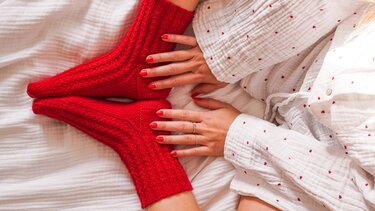 Rote, gestrickte Socken und Hände mit rotem Nagellack auf weißer Bettwäsche mit Herzchen