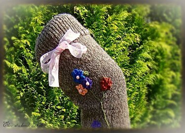 Gestrickte graubraune Socke mit rosa Schleife und blauen, orangefarbenen und roten Häkelblumen vor grünem Laub