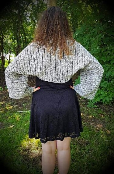 Rückenansicht einer Person mit grau-meliertem, gehäkeltem Shrug mit weiten Ärmeln über schwarzem Kleid vor grünem Hintergrund.