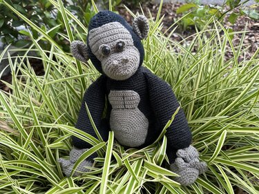 Gehäkelter Amigurumi-Affe in Grau und Dunkelblau sitzt im Ziergras zwischen langen, grün-weiß gestreiften Blättern.
