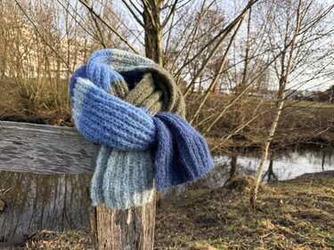 Nahaufnahme eines gestrickten Schals mit Rippenmuster und Farbsegmenten in Blau, Grün und Grau auf Holzpfosten vor Bach