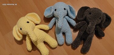 Drei gehäkelte Amigurumi‑Elefanten (gelb, hellblau, dunkelgrau) liegend auf Holzfußboden