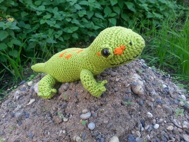 Gehäkeltes hellgrünes Reptil‑Amigurumi mit orangen Punkten und glänzenden Augen auf Erdhügel
