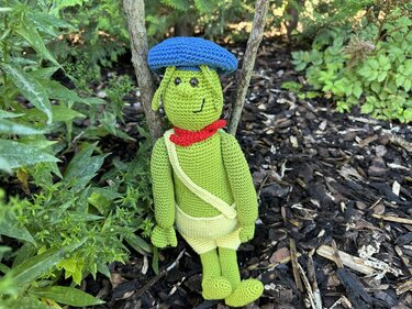 Gehäkelter grüner Amigurumi-Frosch mit blauer Mütze, rotem Halstuch und gelber Hose im Garten
