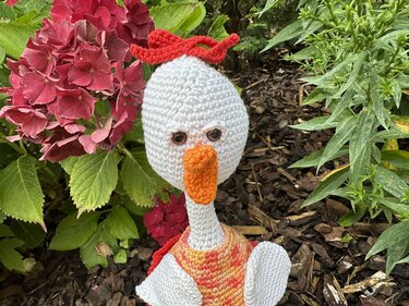 Weiß gehäkelte Amigurumi-Gans mit orange Schnabel und roter Schleife vor Gartenpflanzen