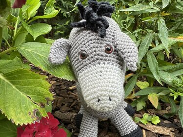 Grau-weißer gehäkelter Amigurumi-Esel mit schwarzer Mähne zwischen grünen Gartenblättern