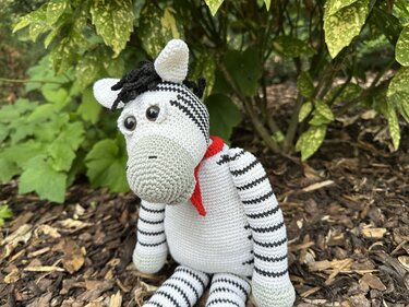 Gehäkeltes Zebra-Amigurumi mit rotem Halstuch auf Rindenmulch vor Pflanzen
