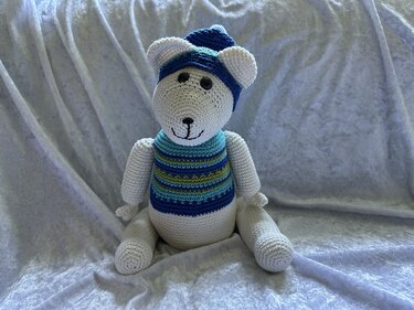 Weißer gehäkelter Amigurumi-Bär mit blauer Mütze und gestreifter Weste auf hellem Stoff