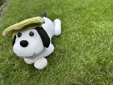 Weißer Amigurumi-Hund mit schwarzen Ohren und grüner Schirmmütze auf Gras