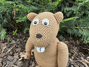 Brauner gehäkelter Amigurumi-Biber mit großen Augen und weißen Nagezähnen vor grünen Zweigen