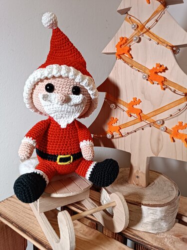 Gehäkelter Amigurumi-Weihnachtsmann mit roter Mütze und weißem Bart auf einem Holzschlitten neben einem dekorativen Holz-Weihnachtsbaum.