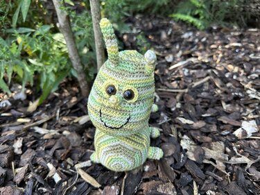 Gehäkelte grün-gelbe Amigurumi-Raupe aus Verlaufsgarn auf Rindenmulch