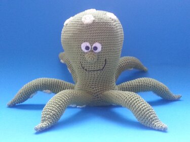 Grüner gehäkelter Amigurumi-Oktopus mit mehreren Armen, weißen Saugnäpfen und lächelndem Gesicht vor blauem Hintergrund