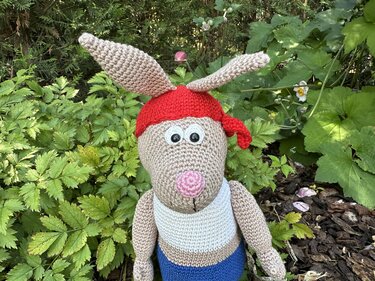 Gehäkelter Amigurumi-Hase mit langen Ohren, rotem Kopftuch, großen Augen, rosa Nase, weißem Oberteil und blauer Hose im Grünen