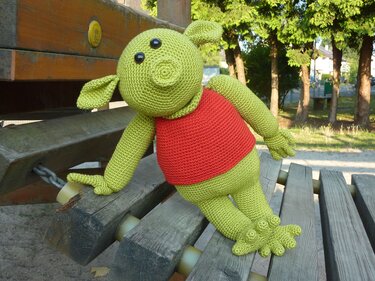 Grünes gehäkeltes Amigurumi mit rotem Shirt sitzt auf einer Holzbank im Park