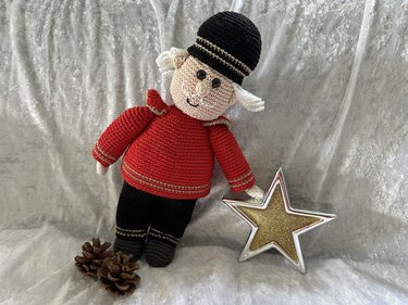 Gehäkelte Nussknacker‑Puppe in roter Uniform und schwarzer Mütze hält goldenen Stern; Tannenzapfen daneben.