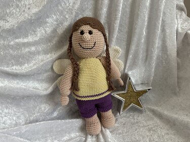 Gehäkelte Amigurumi-Puppe mit braunen Zöpfen, angenähten Flügeln, gelbem Oberteil und violetten Shorts vor silbernem Stoff neben einem Stern