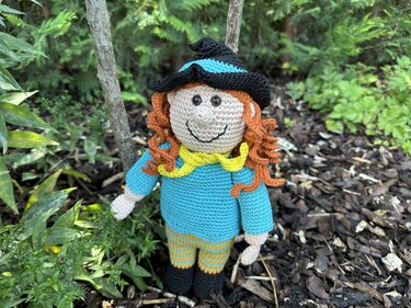 Gehäkelte Hexen-Amigurumi mit schwarzem Hut, türkisfarbenem Kleid und lockigem orangefarbenem Haar im Beet