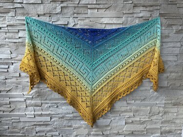 Hand-crocheted triangle shawl in blue, green, and yellow gradient with an open lace pattern and scalloped edge, displayed on a textured wall.