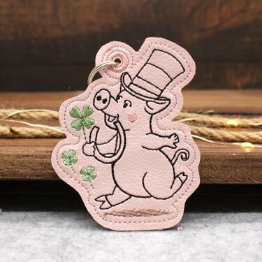 Rosa gestickter Glücksschwein-Schlüsselanhänger mit Zylinder, Hufeisen und Kleeblättern auf Holzuntergrund.