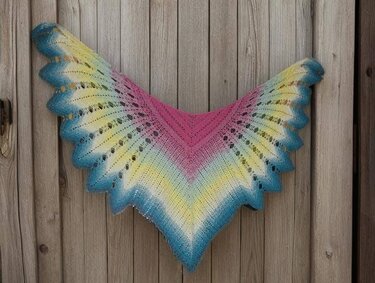 V-shaped crocheted lace shawl in pink, yellow and blue gradient with scalloped edge hanging on a wooden background.